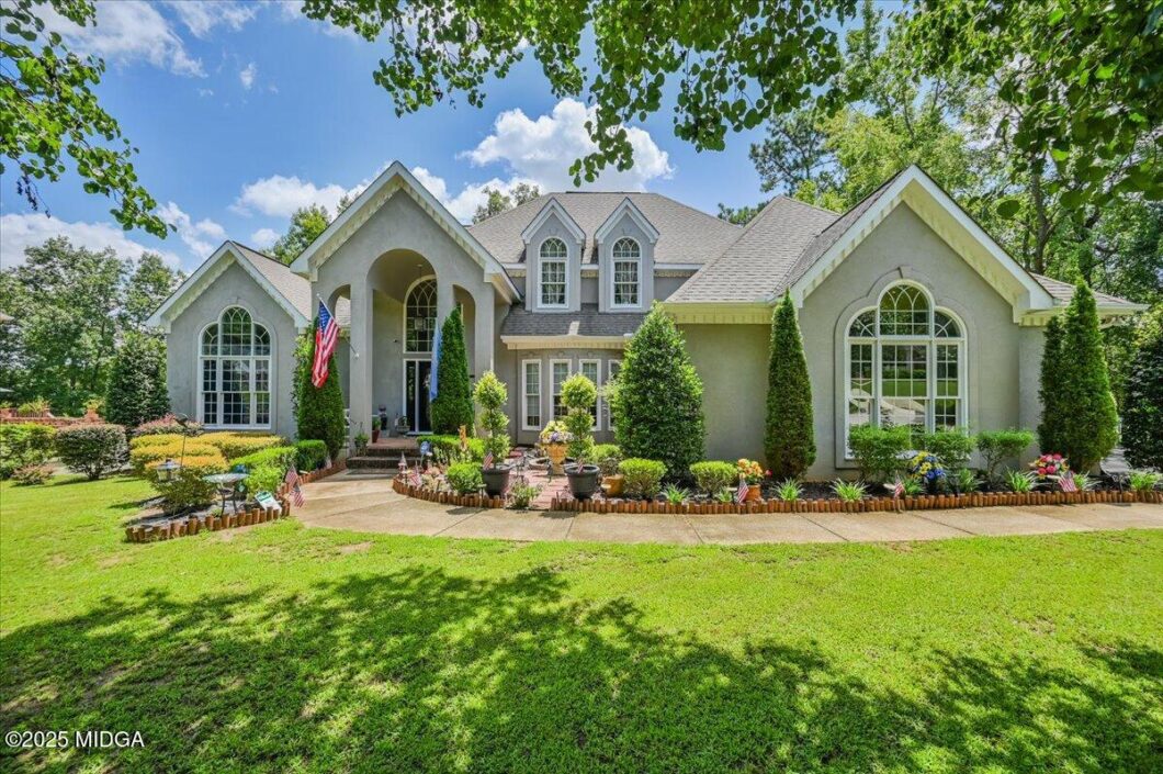 Front view of a gray suburban house with arched windows, a manicured lawn, and a flower-filled border along the walkway, with American flags by the entryway