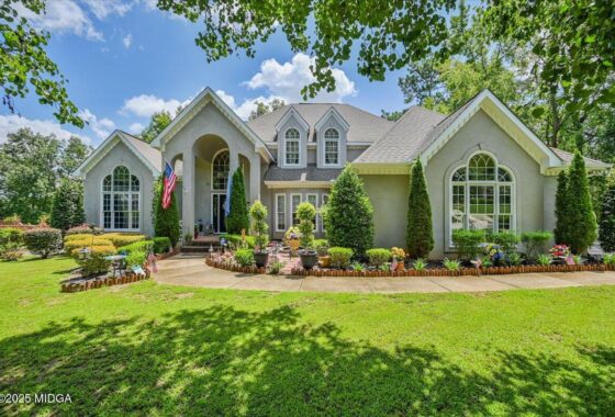 Front view of a gray suburban house with arched windows, a manicured lawn, and a flower-filled border along the walkway, with American flags by the entryway