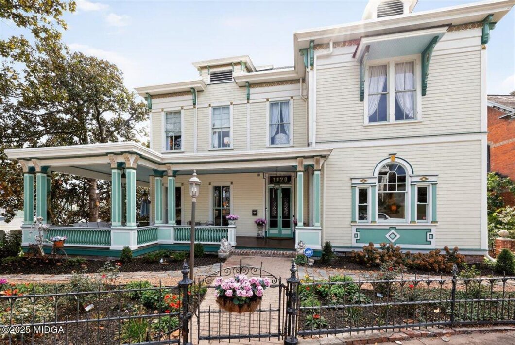 Two-story Victorian house with a wraparound porch, cream siding and teal trim, front garden and wrought-iron fence.