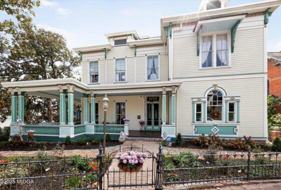 Two-story Victorian house with a wraparound porch, cream siding and teal trim, front garden and wrought-iron fence.