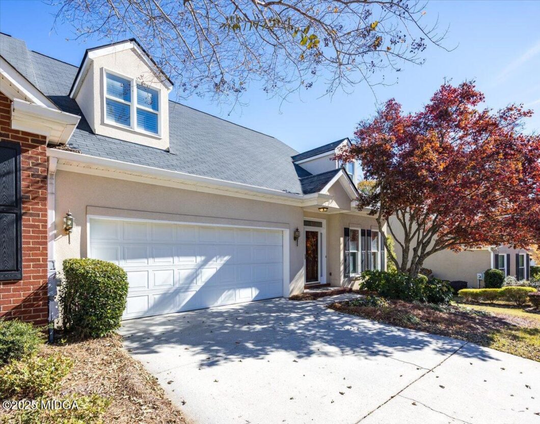 Front view of a beige house with a white two-car garage, dormer window, and a landscaped yard under a sunny blue sky.