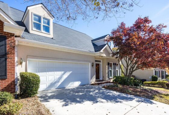 Front view of a beige house with a white two-car garage, dormer window, and a landscaped yard under a sunny blue sky.