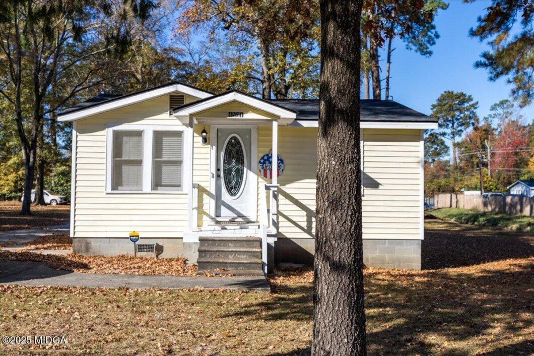 Small beige cottage with white trim, front steps, oval-glass door, and a wreath beside the entry, in autumn sunlight with a large tree in the foreground.