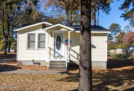 Small beige cottage with white trim, front steps, oval-glass door, and a wreath beside the entry, in autumn sunlight with a large tree in the foreground.