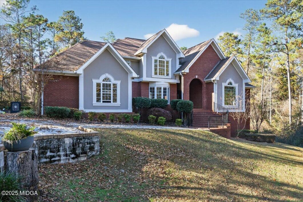 Front view of a two-story brick-and-gray house with arched windows and a curved brick entryway, set on a wooded lot.