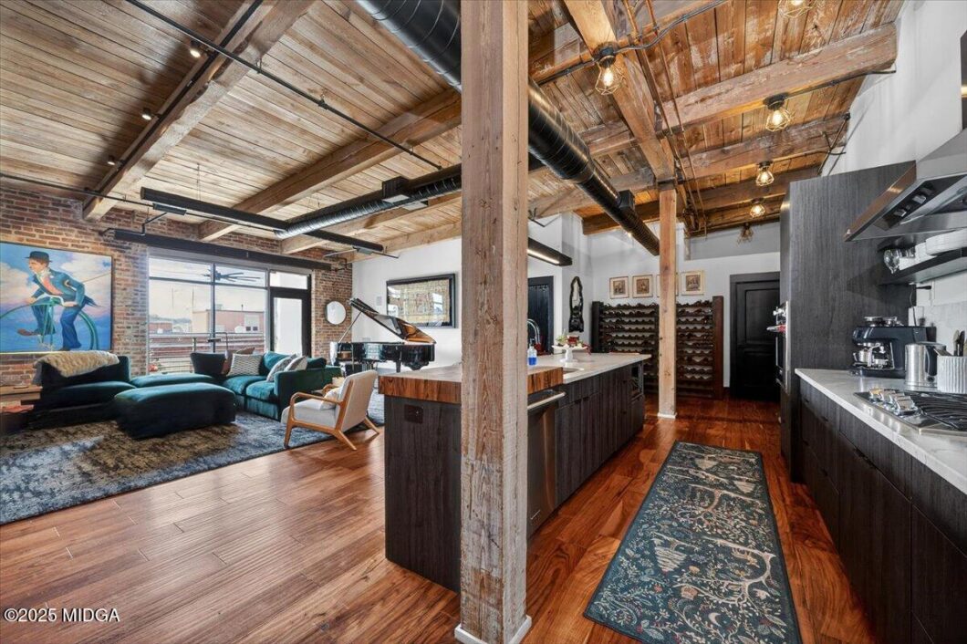 Industrial loft living space with exposed wooden beams, brick wall, and a teal seating area near a grand piano by large windows. A dark wood kitchen island and wine racks are visible in the foreground and right.