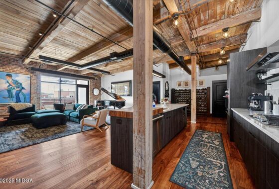 Industrial loft living space with exposed wooden beams, brick wall, and a teal seating area near a grand piano by large windows. A dark wood kitchen island and wine racks are visible in the foreground and right.