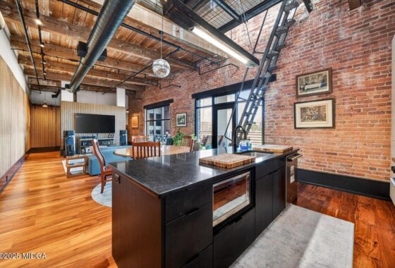 Industrial loft kitchen and living area with brick wall, exposed ducts, dark island, and hardwood floors.