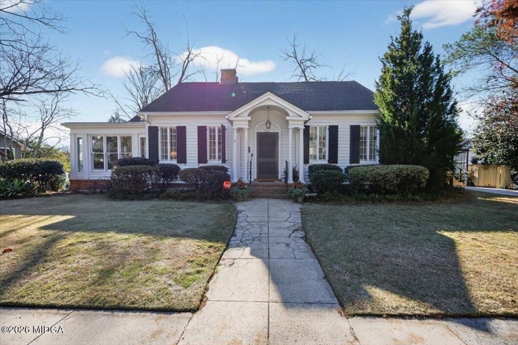 Front view of a single-story white house with black shutters, a central arched entry, and a stone path leading to the front door on a sunny day.