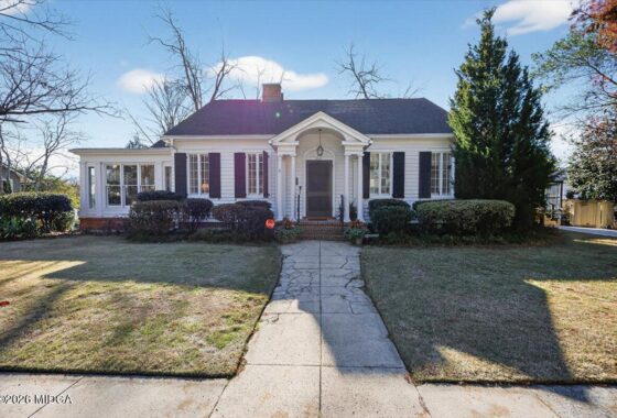 Front view of a single-story white house with black shutters, a central arched entry, and a stone path leading to the front door on a sunny day.