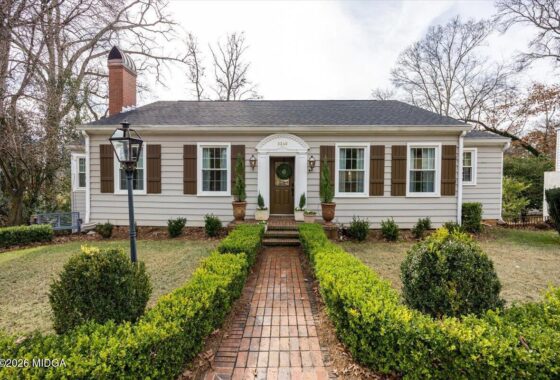 Front view of a single-story beige house with brown shutters, a brick walkway lined by trimmed hedges leading to a centered door with a wreath and planters on each side.