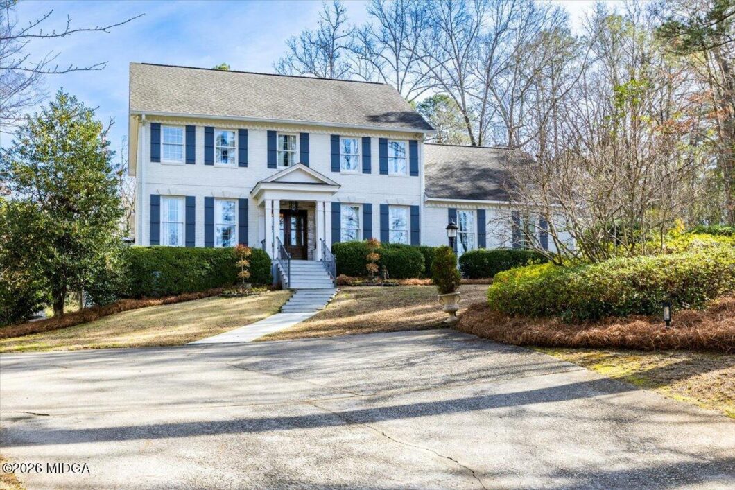 White two-story house with blue shutters, centered front door, and manicured hedges along the entry steps.