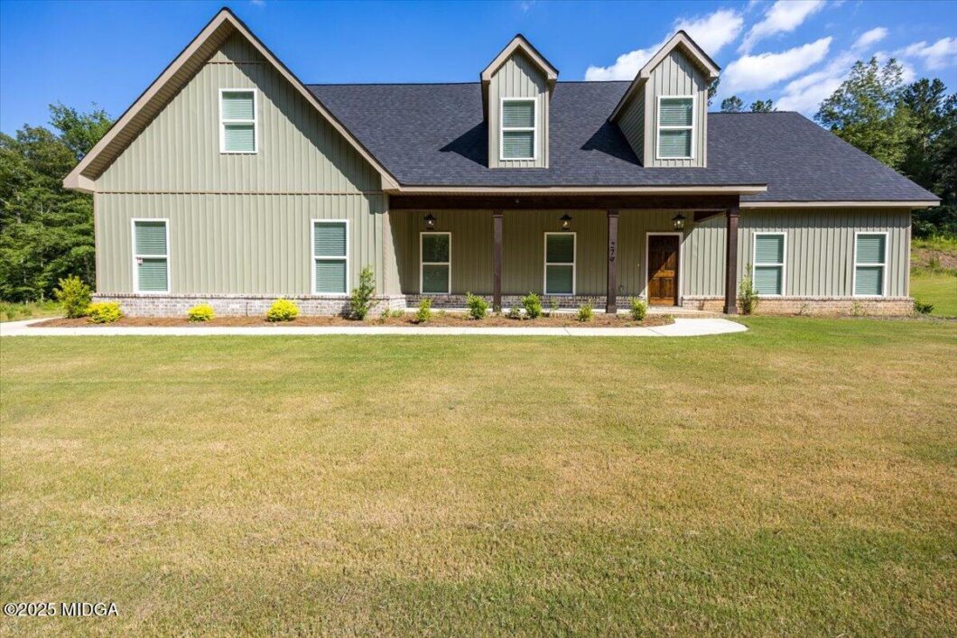 Front view of a two-story suburban house with a covered porch, beige siding, and three dormer windows under a dark roof with a manicured lawn nearby.