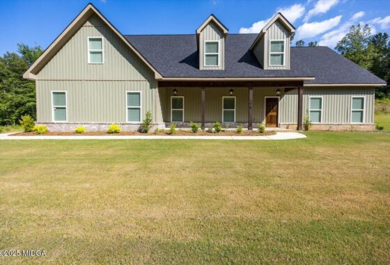 Front view of a two-story suburban house with a covered porch, beige siding, and three dormer windows under a dark roof with a manicured lawn nearby.
