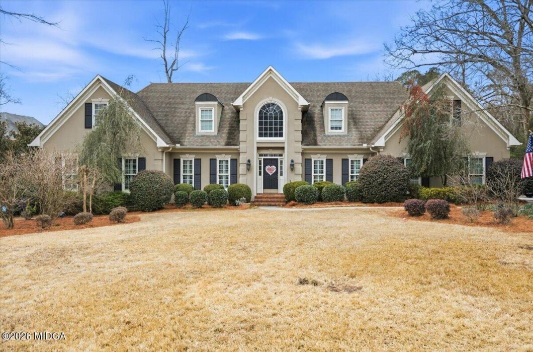 Large two-story suburban home with a centered arched window above the front door, flanked by symmetry and manicured shrubs, under a clear blue sky. A heart wreath hangs on the front door. An American flag is visible on the right side of the yard.
