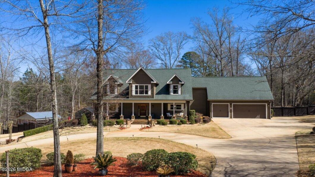 Front view of a two-story house with a green roof, dormer windows, and a covered front porch, plus a three-car garage and curved driveway. Landscaping and leafless trees in a suburban yard.