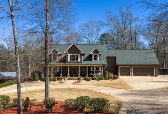 Front view of a two-story house with a green roof, dormer windows, and a covered front porch, plus a three-car garage and curved driveway. Landscaping and leafless trees in a suburban yard.