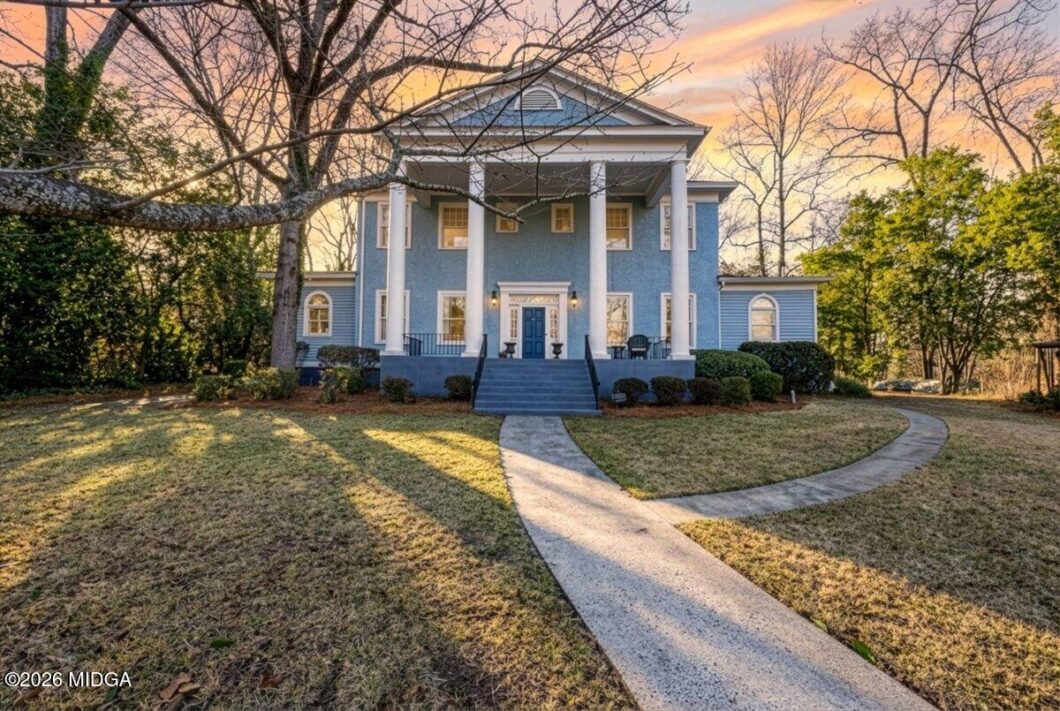 Blue two-story colonial-style home with white columns on a front porch, curved walkway, and bare trees at sunset.