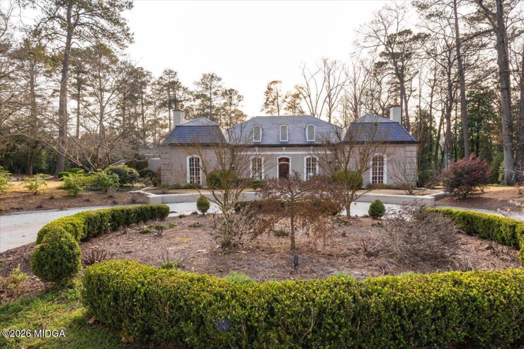 Stately gray manor with dormer windows and slate roof, framed by tall trees, set behind a circular hedge garden.