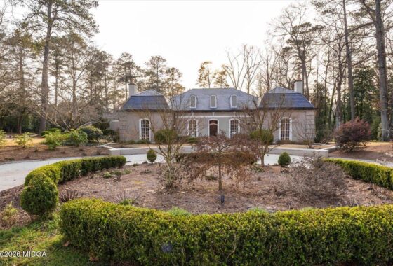 Stately gray manor with dormer windows and slate roof, framed by tall trees, set behind a circular hedge garden.