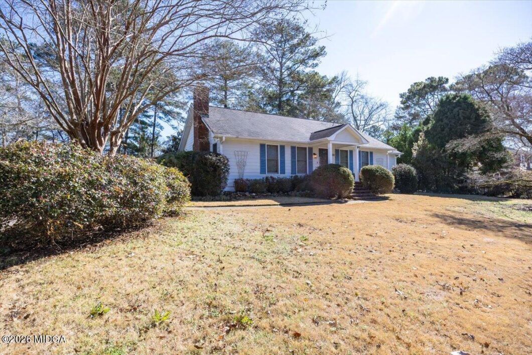 Front view of a single-story blue house with shutters, trimmed bushes, and bare trees on a sunny day.