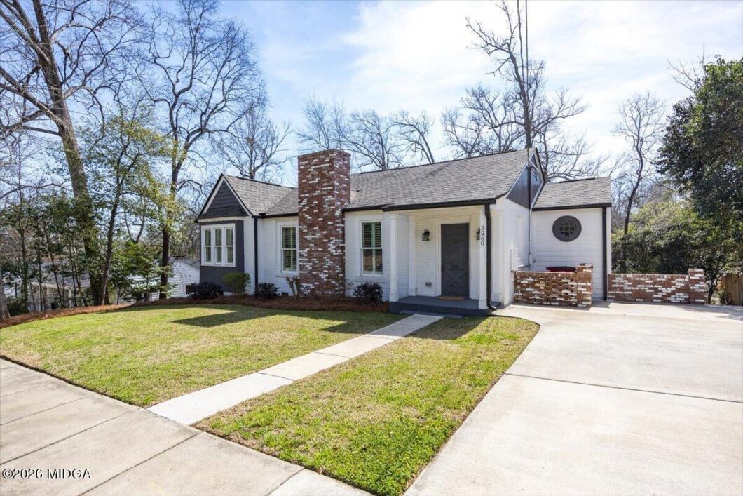 A white one-story house with a brick chimney, gray front door, and a curved concrete walkway leading from the driveway to the porch amid a green lawn and leafless trees.