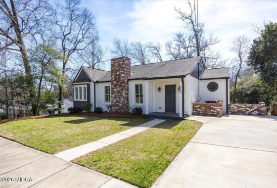 White ranch-style house with a brick chimney, white siding, and a sloped front lawn, with a concrete pathway to the entrance.