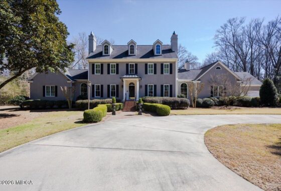 Front view of a large gray, Colonial-style house with dormer windows, black shutters, and a curved driveway lined by trimmed hedges