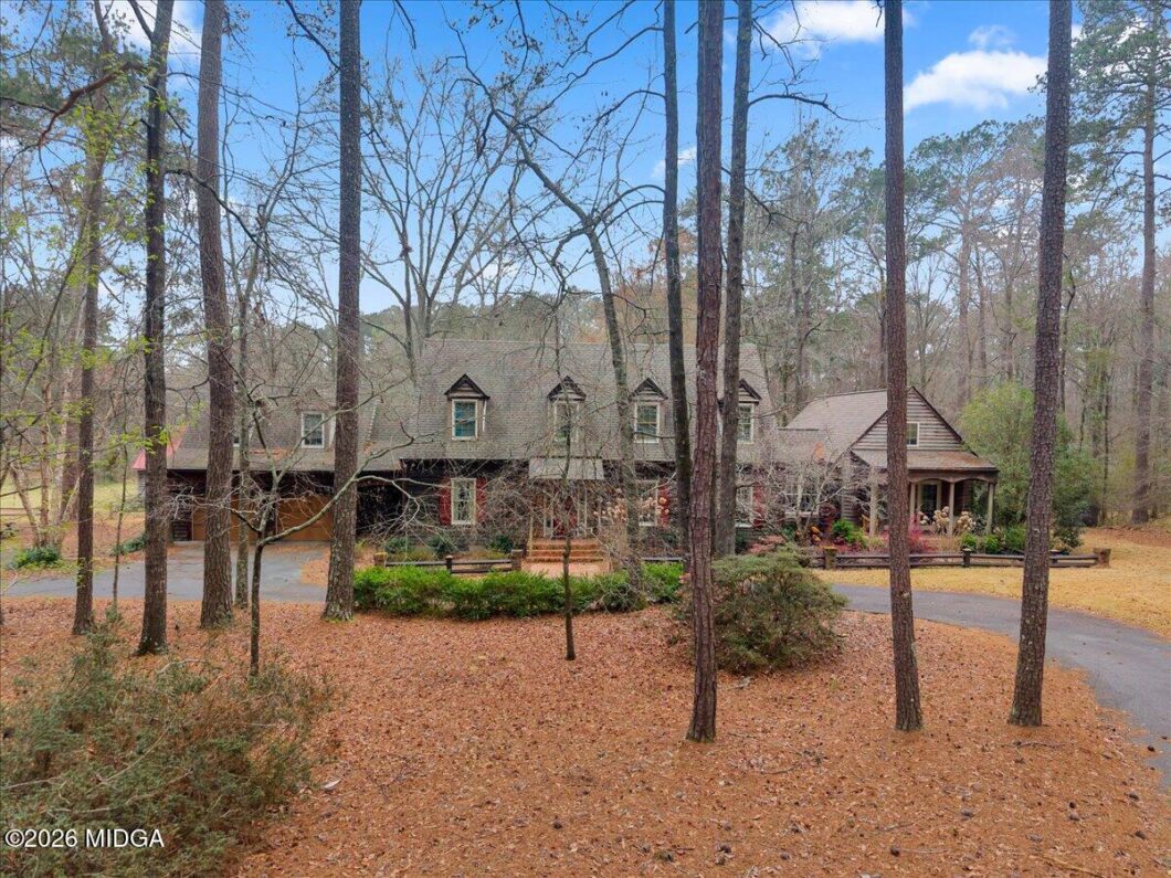 Two-story house with dormers in a pine forest, with a curved driveway and a covered front porch.