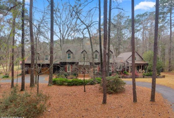 Two-story house with dormers in a pine forest, with a curved driveway and a covered front porch.