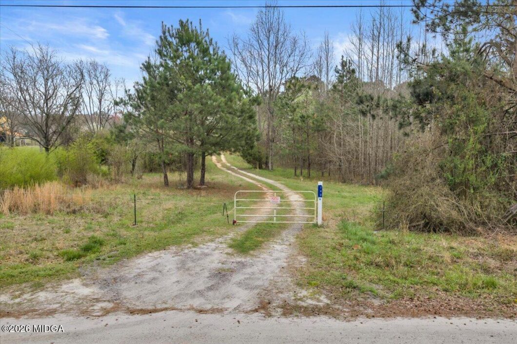 Metal gate blocks a dirt path winding into a tree-filled field.”, “Closed white gate across a country dirt road with trees on both sides.”, “Gated driveway leading into a wooded area, with grass and pines nearby.”, “Rural path blocked by a gate; grass, trees, and a utility post in view.”, “Gate blocking access to a gravel road that leads into a forested area.”