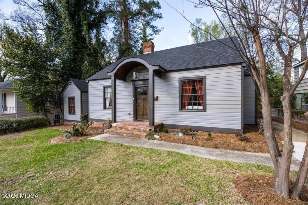 Front view of a small grey suburban house with a curved portico, brick steps, and a well-kept yard with trees nearby