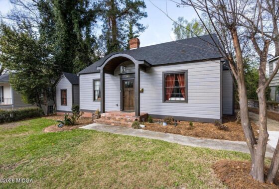 Front view of a small grey suburban house with a curved portico, brick steps, and a well-kept yard with trees nearby