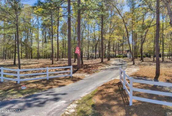 Paved driveway winds through a pine forest toward a single-story house, with white split-rail fences and an American flag on a tree.