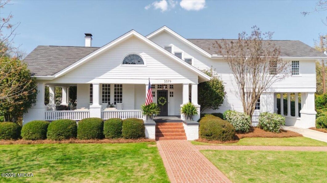 White two-story house with a front porch, gabled roof, and an American flag by the entrance; brick walkway leads to brick steps with potted plants on the porch.