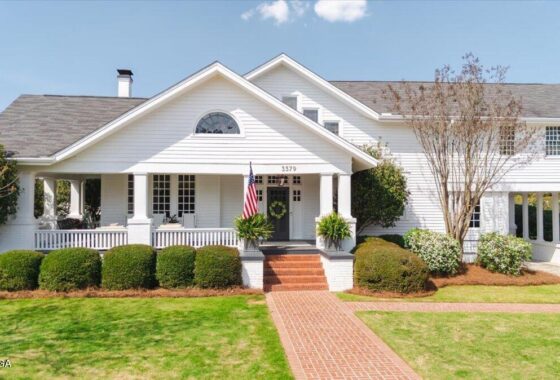 White two-story house with a front porch, gabled roof, and an American flag by the entrance; brick walkway leads to brick steps with potted plants on the porch.