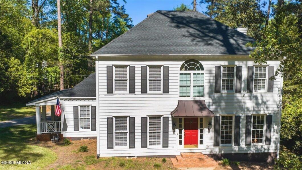 Front view of a white two-story house with black shutters, a red front door, brick steps, and a small porch with an American flag to the left, surrounded by trees.