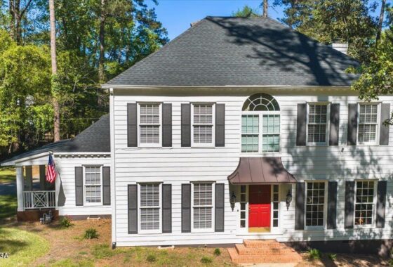 Front view of a white two-story house with black shutters, a red front door, brick steps, and a small porch with an American flag to the left, surrounded by trees.