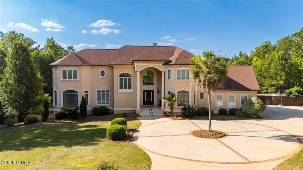 Two-story beige house with a brown roof, arched entry, and circular paved driveway with a central island and palm tree.