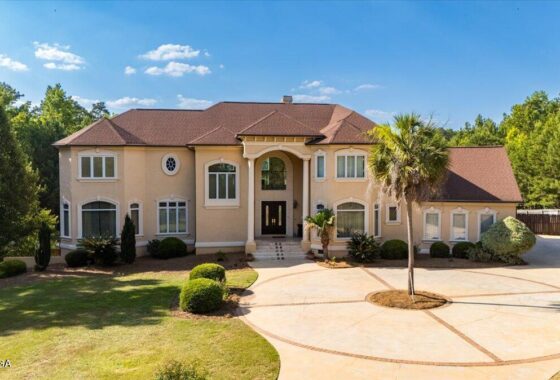 Two-story beige house with a brown roof, arched entry, and circular paved driveway with a central island and palm tree.