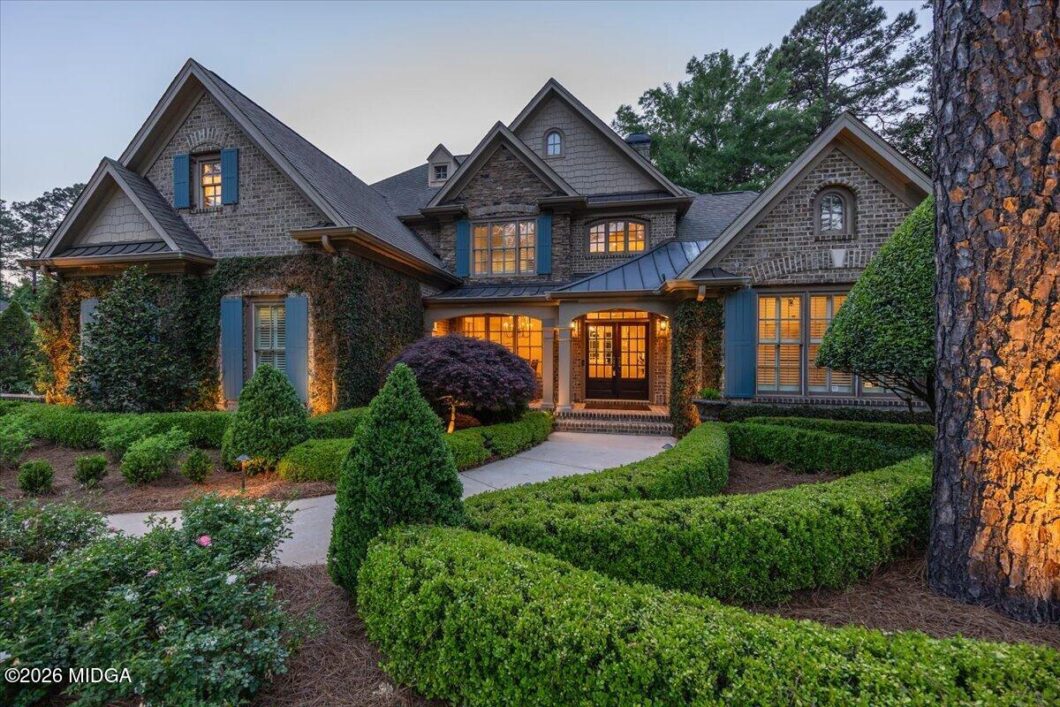 Front view of a large stone-and-brick house with blue shutters, ivy-covered walls, and manicured hedges at dusk, warm lights in windows.