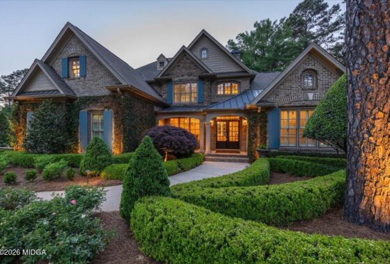 Front view of a large stone-and-brick house with blue shutters, ivy-covered walls, and manicured hedges at dusk, warm lights in windows.