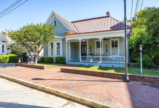 Blue Victorian-style house with a white railing porch on a brick sidewalk, with a tree and street lamp nearby on a sunny day.