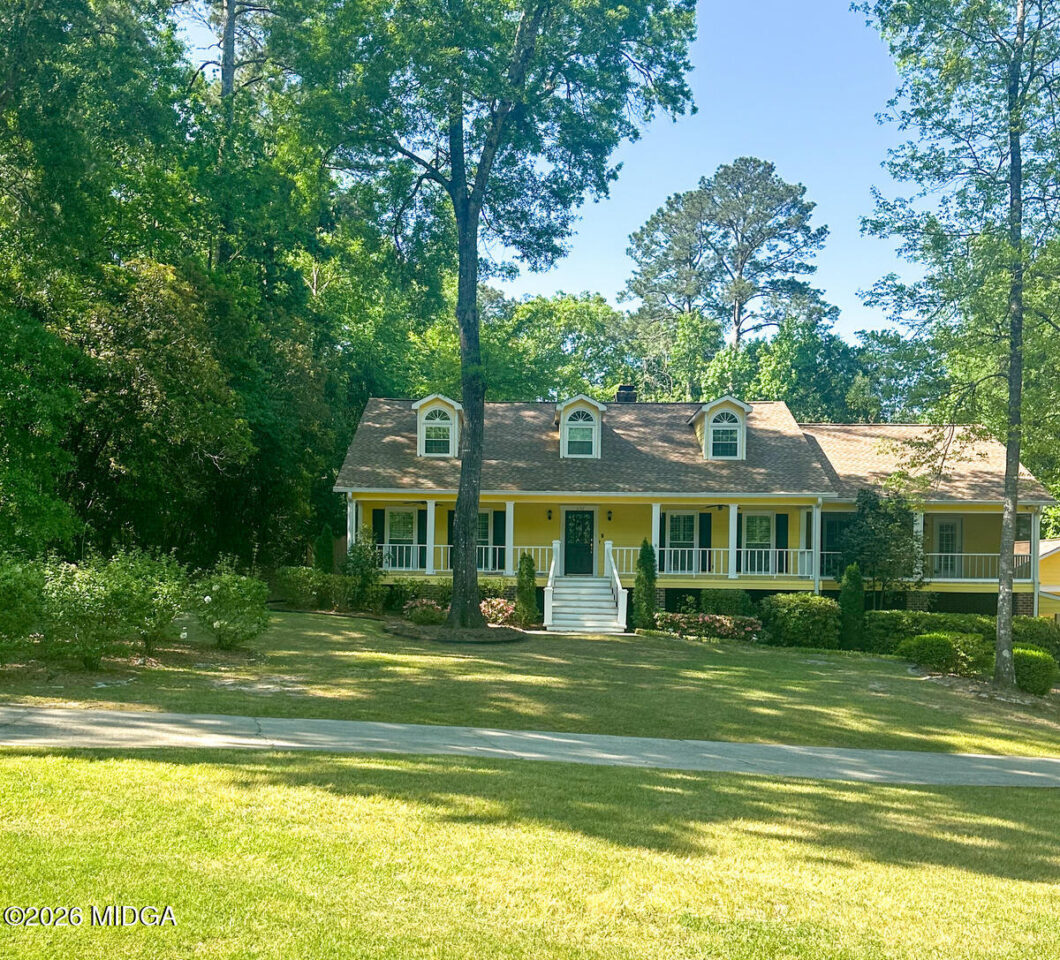 Yellow, single-story house with a white wraparound porch, three dormer windows, and lush trees framing the front yard on a sunny day