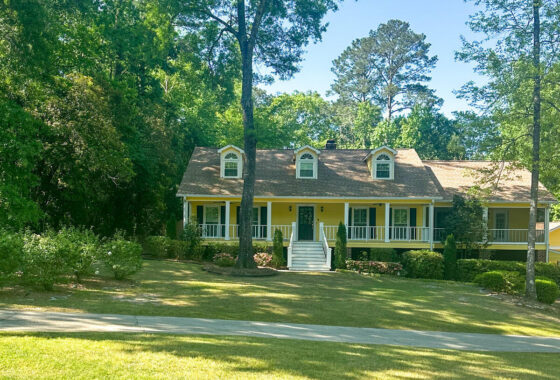 Yellow, single-story house with a white wraparound porch, three dormer windows, and lush trees framing the front yard on a sunny day