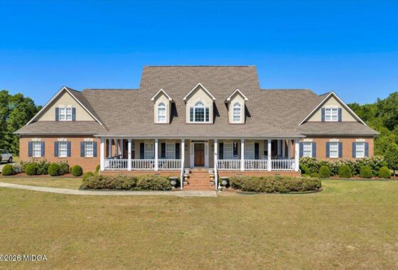 Front view of a large brick suburban home with a wraparound white porch, dormer windows, and a manicured lawn on a sunny day.