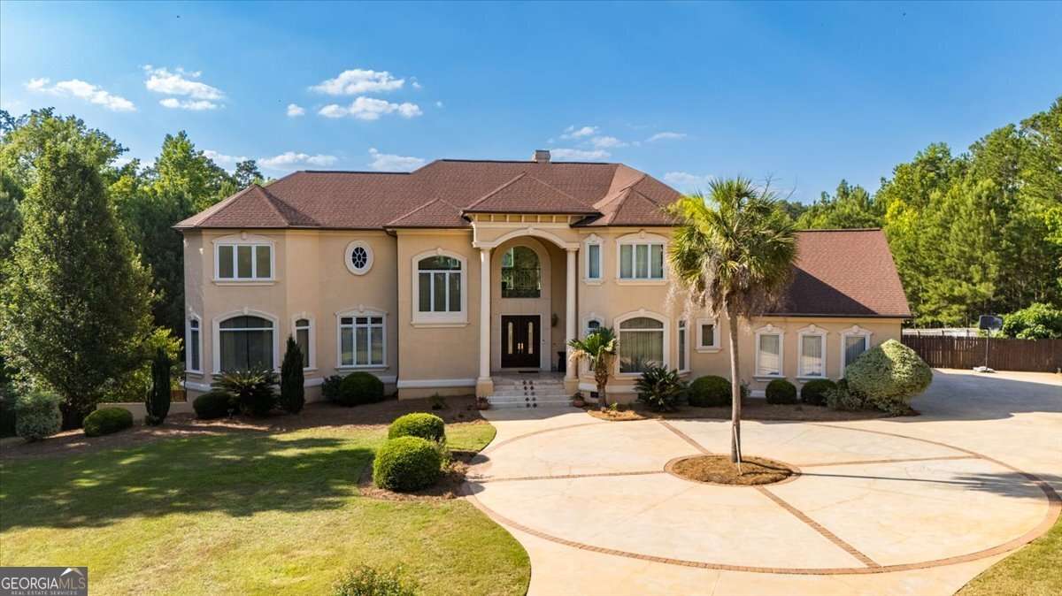 Front view of a two-story beige house with a brown roof, circular brick driveway, and well-kept landscaping including a palm tree and shrubs.