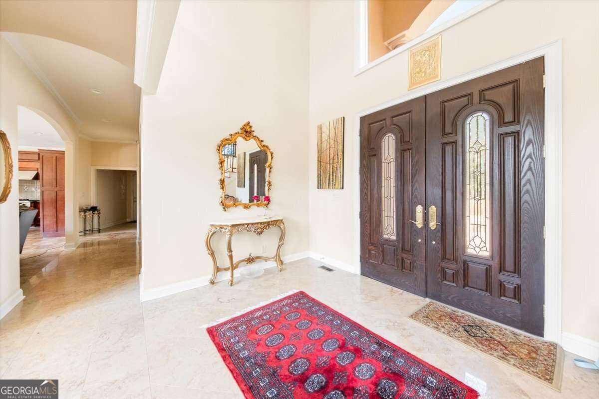 Elegant foyer with dark wooden double doors, gold-framed mirror, and marble floor; red patterned runner rug and decorative console table nearby.