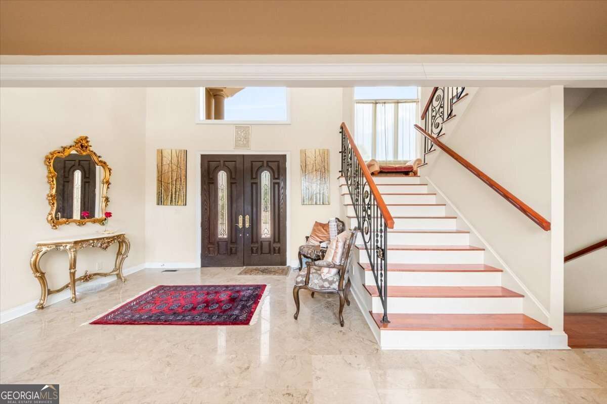 Elegant foyer with marble floor, gold-framed mirror above a gilded console table, dark double doors, a red patterned rug, and a curved staircase with wooden handrails on the right.