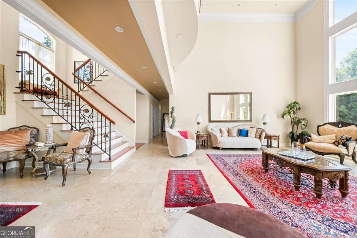 Bright living room with cream sofas, ornate coffee table, and a curved staircase with iron railing under a high ceiling.